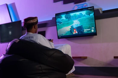 الجلوس على المقعد الاسود الاسفنجي المريح اثناء اللعب داخل مركز الالعاب الالكترونية , رجل عربي خليجي عماني يرتدي الدشداشة البيضاء و العمامة يحمل جهاز التحكم عن بعد , النظر الي الشاشة المعلقة على الحائط