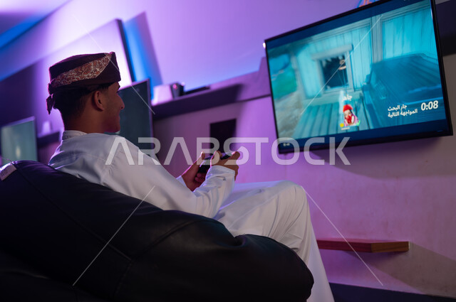 التحكم بسهولة فب اللعبة عن طريق جهاز التحكم اللاسلكية , رجل عربي خليجي عماني يرتدي الدشداشة البيضاء و العمامة يجلس على مقعد اسفنجي اسود , ايماءات السعادة و الابتسامة