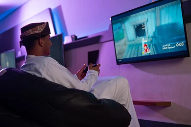 التحكم بسهولة فب اللعبة عن طريق جهاز التحكم اللاسلكية , رجل عربي خليجي عماني يرتدي الدشداشة البيضاء و العمامة يجلس على مقعد اسفنجي اسود , ايماءات السعادة و الابتسامة