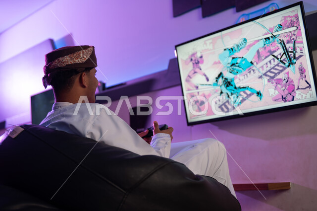 النظر الي الشاشة ذات الالوان البراقة , رجل عربي خليجي عماني يرتدي الدشداشة البيضاء و العمامة يلعب منفرداً بتركيز داخل مركز الالعاب الالكترونية , صورة خلفية