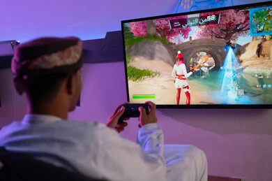 شاشة تلفزيون معلقة على الحائط تعرض لعبة الكترونية ذات الوان زاهية , رجل عربي خليجي عماني يرتدي دشداشة بيضاء و عمامة بنية يحمل جهاز التحكم عن بعد اللاسلكي لسهولة الحركة , التركيز اثناء اللعب , صورة خلفية