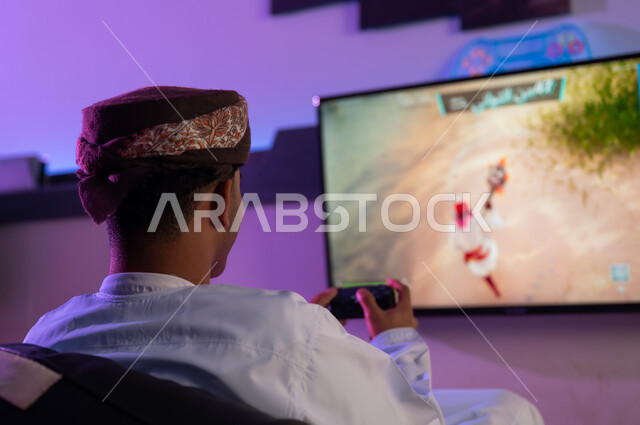 صورة خلفية لرجل عربي خليجي عماني يرتدي الدشداشة البيضاء و العمامة يجلس على كرسي اسود اسفنجي , النظر الي الشاشة المعلقة على الحائط , اللعب بتركيز و دقة