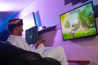 انشطة شبابية فردية مسلية , رجل عربي خليجي عماني يرتدي الدشداشة البيضاء و العمامة يحمل جهاز التحكم عن بعد باللعبة الالكترونية , النظر للشاشة المعلقة على الحائط بسعادة