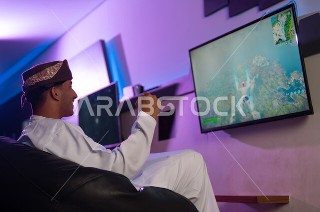 Raising a fist in the air as a sign of victory in the electronic game, an Arab Gulf Omani man wearing a turban and a white dishdasha sits in front of the screen, gestures of joy and happiness at winning.