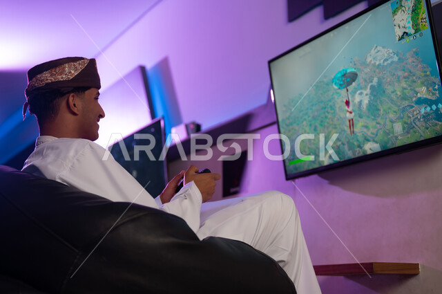 النظر الي الشاشة المعلقة على الحائط , رجل عربي خليجي عماني يرتدي عمامة بنية و دشداشة بيضاء يحمل جهاز التحكم عن بعد باللعبة الالكترونية , صورة جانبية