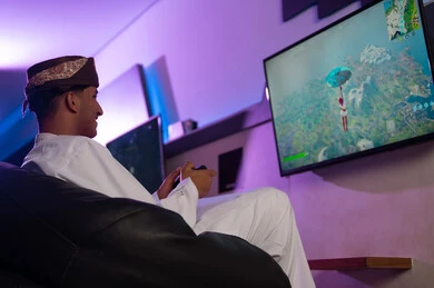 النظر الي الشاشة المعلقة على الحائط , رجل عربي خليجي عماني يرتدي عمامة بنية و دشداشة بيضاء يحمل جهاز التحكم عن بعد باللعبة الالكترونية , صورة جانبية