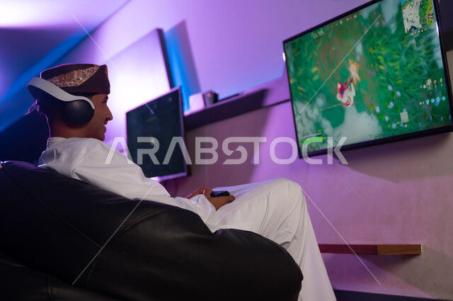 التواصل الحي مع الاصدقاء في الالعاب الالكترونية باستخدام خدمات الانترنت , رجل عربي خليجي عماني يرتدي الدشداشة البيضاء و العمامة يجلس على الكرسي المريح , انشطة شبابية جماعية و فردية