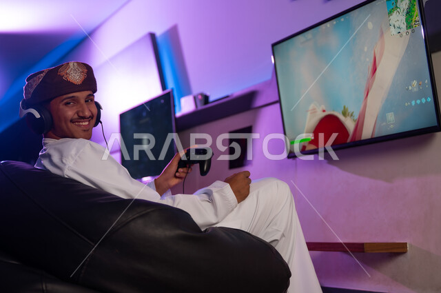 شاب عربي خليجي عماني يرتدي دشداشة بيضاء و عمامة بنية يجلس على كرسي اسود اسفنجي مريح , مركز العاب الكترونية , النظر الي الكاميرا بابتسامة و سعادة ,