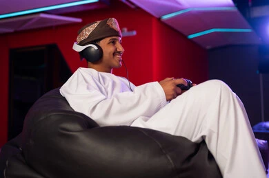 النظر الي الشاشة على الحائط , رجل عربي خليجي عماني يرتدي العمامة و الدشداشة البيضاء يجلس على كرسي اسفنجي اسود , اللعب بسعادة بالتواصل الحي بين الاصدقاء باستخدام سماعة الرأس