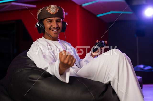 قفل قبضة اليد ورفعها للاعلى علامة على الفوز و الحماس , رجل عربي خليجي عماني يرتدي الدشداشة البيضاء و العمامة البنية يجلس على كرسي اسود اسفنجي , وضع سماعة الرأس للتواصل المباشر مع الاصدقاء , ايماءات السعادة و الفرحة