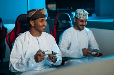 Looking at the screen with happiness while playing electronic football, two Arab Gulf men from Oman wearing white dishdasha and brown turban, with embroidered kumma, holding the remote control, gestures of happiness and joy.