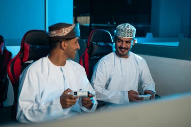 اوقات المتعة و التسلية بين الاصدقاء داخل مركز الالعاب الالكترونية , رجلان عربيان خليجيان عمانيان يرتديان الدشداشة البيضاء و العمامة البنية , الكمة المطرزة , النظر بسعادة قبل بداية اللعبة
