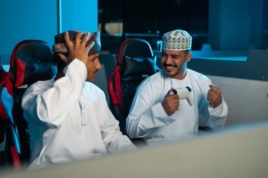 A Gulf Arab Omani man wearing a turban and a white dishdasha places his hand on his head as a sign of loss and deep sadness, while a young Gulf Arab Omani man wearing a white dishdasha and an embroidered kumma celebrates winning in the electronic game.