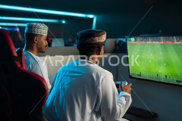 شاشة تلفزيون تعرض لعبة كرة القدم الالكترونية , رجلان عربيان خليجيان عمانيان يرتديان دشداشة بيضاء و عمامة بنية , الكمة المطرزة , لعبة تنافسية مسلية بين الاصدقاء