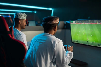 شاشة تلفزيون تعرض لعبة كرة القدم الالكترونية , رجلان عربيان خليجيان عمانيان يرتديان دشداشة بيضاء و عمامة بنية , الكمة المطرزة , لعبة تنافسية مسلية بين الاصدقاء