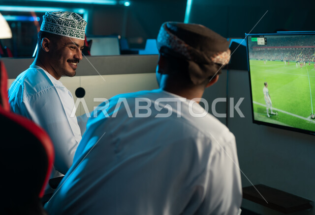 حديث ممتع بين الاصدقاء اثناء اللعب داخل مركز الالعاب الالكتروني , رجلان عربيان خليجيان عمانيان يرتديان دشداشة بيضاء و عمامة , الكمة المطرزة , ايماءات سعادة و بهجة , انشطة شبابية مسلية