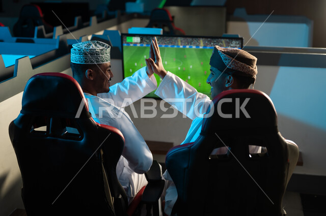 A handshake of the palms raised as a sign of victory in the game, two Arab Gulf men from Oman wearing white dishdasha and embroidered kumma, a brown turban, gestures of happiness and joy at winning the electronic game.