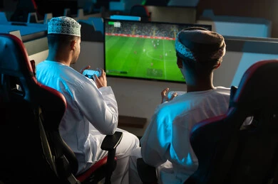 النظر بحماسة الي شاشة تعرض لعبة كرة القدم الالكترونية , رجلان عربيان خليجيان يرتديان الدشداشة البيضاء و العمامة البنية و الكمة المطرزة يجلسان داخل مركز الالعاب الالكترونية