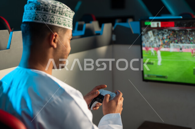 النظر الي شاشة تعرض لعبة كرة القدم , رجل عربي خليجي عماني يرتدي الدشداشة البيضاء و الكمة المطرزة يحمل جهاز التحكم عن بعد في اللعبة الالكترونية , لعبة حماسية ممتعة