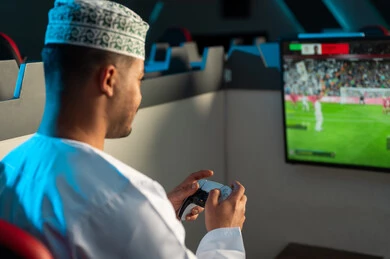 النظر الي شاشة تعرض لعبة كرة القدم , رجل عربي خليجي عماني يرتدي الدشداشة البيضاء و الكمة المطرزة يحمل جهاز التحكم عن بعد في اللعبة الالكترونية , لعبة حماسية ممتعة