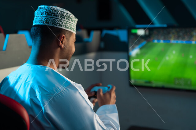 صورة خلفية لرجل خليجي عماني عربي يرتدي الدشداشة البيضاء و الكمة المطرزة ينظر الي شاشة معلقة تعرض لعبة كرة القدم , ايماءات السعادة و الفرحة اثناء اللعب
