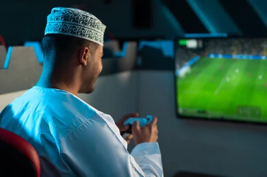 A background image of a Gulf Omani Arab man wearing a white dishdasha and an embroidered kumma, looking at a hanging screen displaying a football game, with gestures of happiness and joy while playing.