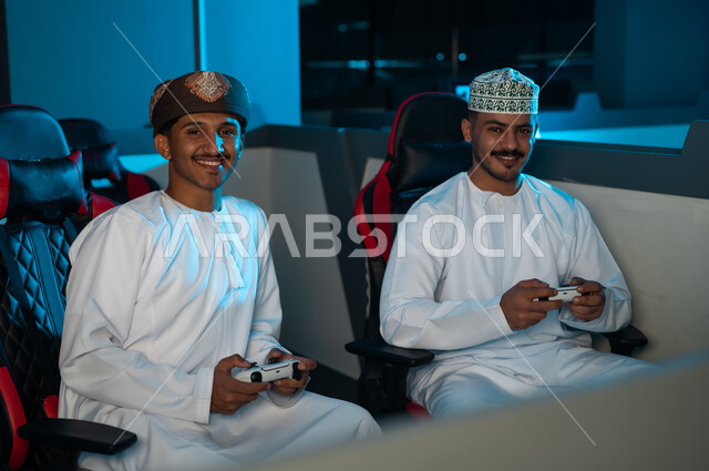 النظر الى الكاميرا بسعادة , رجلان عربيان خليجان عمانين يرتديان الدشداشة البيضاء و الكمة المطرزة , عمامة لون بني , الامساك بجهاز التحكم عن بعد للعبة الالكترونية , ايماءات الفرح بمشاركة الاوقات السعيدة مع الاصدقاء