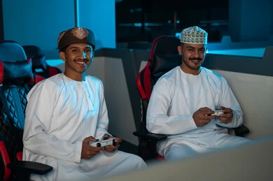 النظر الى الكاميرا بسعادة , رجلان عربيان خليجان عمانين يرتديان الدشداشة البيضاء و الكمة المطرزة , عمامة لون بني , الامساك بجهاز التحكم عن بعد للعبة الالكترونية , ايماءات الفرح بمشاركة الاوقات السعيدة مع الاصدقاء