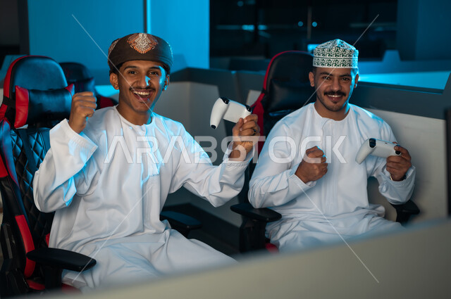 رجلان عربيان خليجيان عمانيان يرتديان الدشداشة البيضاء و الكمة المطرزة و العمامة البنية , رفع اليدين في الهواء اشارة الي الفرح الشديد بعد الفوز في اللعبة الالكترونية , ايماءات السعادة و الفرحة