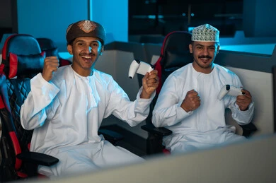 رجلان عربيان خليجيان عمانيان يرتديان الدشداشة البيضاء و الكمة المطرزة و العمامة البنية , رفع اليدين في الهواء اشارة الي الفرح الشديد بعد الفوز في اللعبة الالكترونية , ايماءات السعادة و الفرحة