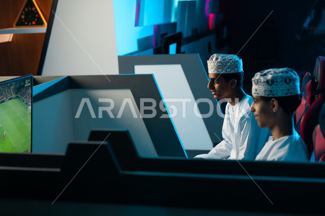 Two Arab Gulf young men from Oman, wearing white dishdasha and embroidered kumma, are sitting inside the private room in the electronic games center, looking at the television screen mounted on the wall, playing the famous electronic football game.