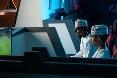 شابان عربيان خليجيان عمانيان يرتديان دشداشة بيضاء و كمة مطرزة يجلسان داخل الغرفة الخاصة في مركز الالعاب الالكترونية , النظر الي شاشة التلفزيون المعلقة على الحائط , لعبة كرة القدم الالكترونية االشهيرة