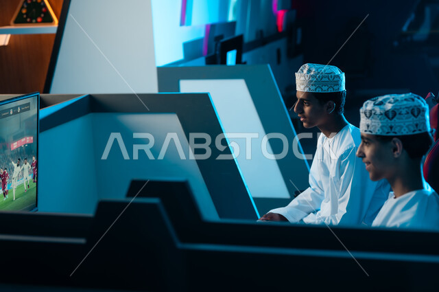 النظر للشاشة المعلقة على الحائط , شابان عربيان خليجيان عمانيان يرتديان دشداشة بيضاء و كمة مطرزة يجلسان على مقاعد جلدية مريحة , اللعب بتركيز مع الاصدقاء