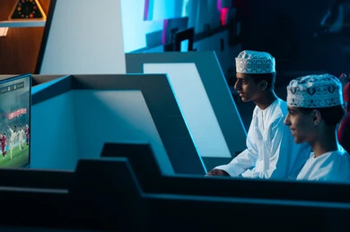 النظر للشاشة المعلقة على الحائط , شابان عربيان خليجيان عمانيان يرتديان دشداشة بيضاء و كمة مطرزة يجلسان على مقاعد جلدية مريحة , اللعب بتركيز مع الاصدقاء