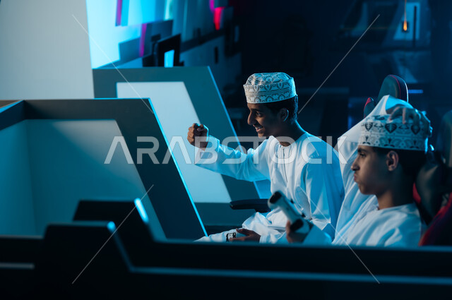 A young Arab Gulf Omani man places his hand on his head in sadness and regret over the loss, while raising his fist in the air as a sign of victory over his friend. Expressions of happiness from winning, and expressions of sadness from losing in the electronic game.