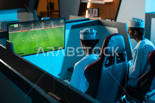 The famous electronic football game beloved by young people, two Arab Gulf young men from Oman wearing white dishdasha and embroidered kumma are looking at the hanging screen, sharing happy moments with friends inside electronic gaming centers.