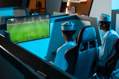 لعبة كرة القدم الالكترونية الشهيرة المحببة للشباب , شابان عربيان خليجيان عمانيان يرتديان الدشداشة البيضاء و الكمة المطرزة ينظران للشاشة المعلقة , مشاركة الاوقات السعيدة مع الاصدقاء داخل مراكز الالعاب الالكترونية