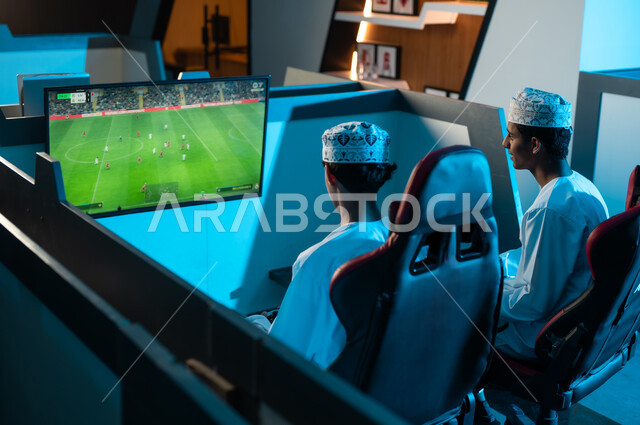 النظر الى الشاشة المعلقة على الحائط , شابان عربيان خليجيان عمانيان يرتديان الدشداشة البيضاء و الكمة المطرزة يجلسان على المقاعد الجلدية المريحة , العاب شبابية حماسية داخل مركز الالعاب الالكترونية