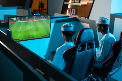 النظر الى الشاشة المعلقة على الحائط , شابان عربيان خليجيان عمانيان يرتديان الدشداشة البيضاء و الكمة المطرزة يجلسان على المقاعد الجلدية المريحة , العاب شبابية حماسية داخل مركز الالعاب الالكترونية