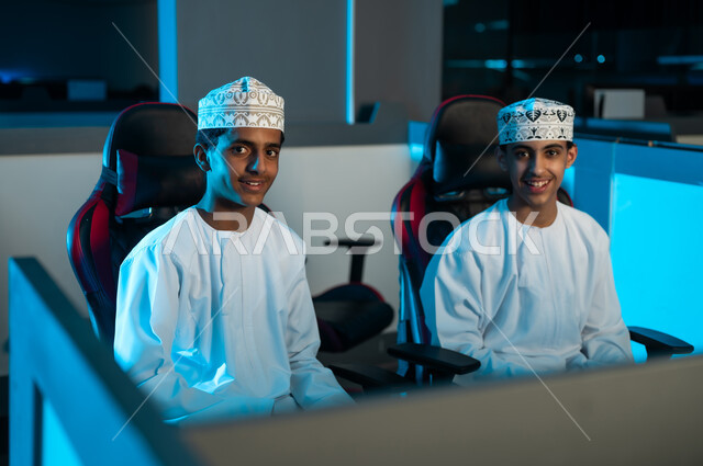 شابان عربيان خليجيان عمانيان يرتديان الدشداشة البيضاء و الكمة المطرزة ينظران للكاميرا في سعادة , ايماءات الفرحة داخل مركز الالعاب الالكترونية , الجلوس على المقاعد الجلدية المريحة