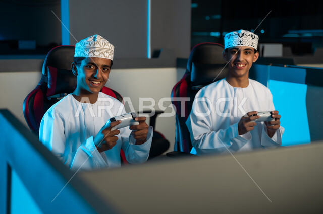 الامساك بجهاز التحكم في اللعبة الالكترونية عن بعد , شابان عربيان خليجيان عمانيان يرتديان الدشداشة البيضاء و الكمةالمطرزة يجلسان على مقاعد جلدية , النظر للكاميرا , ايماءات السعادة و الفرحة قبل بداية اللعب مع الاصدقاء