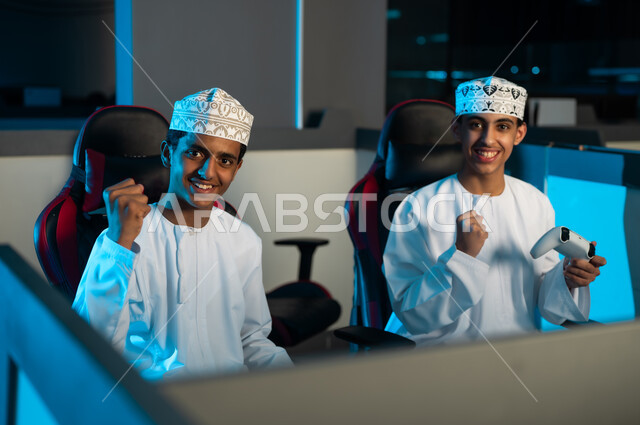 النظر للكاميرا باتسامة بعد تحقيق الفوز , شابان عربيان خليجيان عمانيان يرتديان الدشدشاة البيضاء و الكمة المطرزة يرفعان قبضة اليد لاعلى اشارة على النصر , العاب تنافسية شبابية ممتعة , ايماءات الفرحة و السعادة