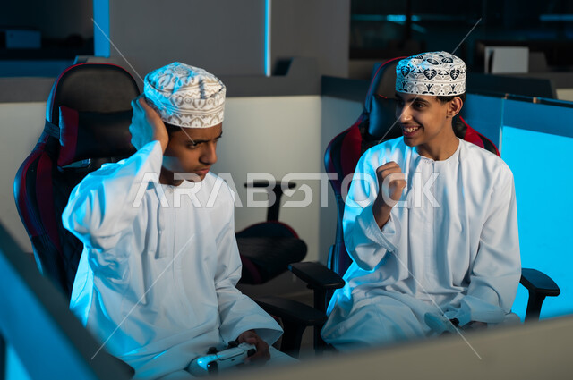 شاب عربي خليجي عماني يرتدي الدشداشة البيضاء و الكمة المطرزة يقفل قبضة اليد و يرفعها لاعلى علامة على الفوز , الصديق يشعر بالحزن للخسارة امام صديقه , العاب المنافسة بين الشباب , الاوقات المسلية الممتعة