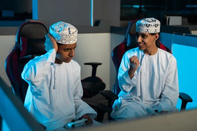 شاب عربي خليجي عماني يرتدي الدشداشة البيضاء و الكمة المطرزة يقفل قبضة اليد و يرفعها لاعلى علامة على الفوز , الصديق يشعر بالحزن للخسارة امام صديقه , العاب المنافسة بين الشباب , الاوقات المسلية الممتعة