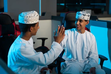 شابان عربيان عمانيان يرتديان الدشداشة البيضاء و الكمة المطرزة يضربان الكفين ببعضهما عالياً اشارة على تحقيق الفوز في اللعبة الالكترونية , الجلوس على المقاعد الجلدية داخل مركز الالعاب الالكترونية , ايماءات السعادة و الفرحة بالنصر