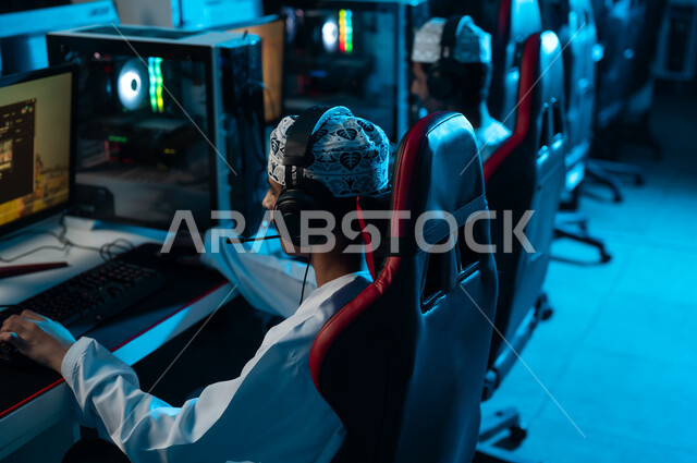 لعب الالعاب الالكترونية الجماعية عن بعد باستخدام خدمات الانترنت , شابان عربيان خليجيان عمانيان يرتديان دشداشة بيضاء و كمة مطرزة يجلسان امام شاشة الحاسب المكتبي , التواصل مع اللاعبيين المشاركين في اللعبة بسهولة