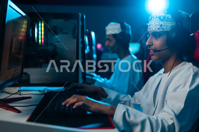 حديث مباشر بين اللاعبين المشاركين في اللعبة الالكترونية باستخدام سماعة الرأس , شابان عربيان خليجيان عمانيان يرتديان دشداشة بيضاء و كمة مطرزة ينظران الى الشاشة بتركيز , ايماءات السعادة و الفرحة