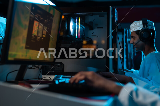 عرض اللعبة الالكترونية على شاشة الحاسب المكتبي , شاب عربي خليجي عماني يرتدي الدشداشة البيضاء و الكمة المطرزة يجلس بجانب صديقه , النقر على لوحة المفاتيح , ايماءات سعادة , صورة مقربة ,