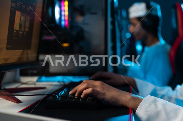 النقر على لوحة مفاتيح الحاسب المكتبي , شاب عربي خليجي عماني يرتدي الدشداشة البيضاء و الكمة المطرزة يجلس على المقعد الجلدي الاحمر , النظر الي شاشة تعرض اللعبة الالكترونية , مشاركة الاصدقاء الاوقات المسلية