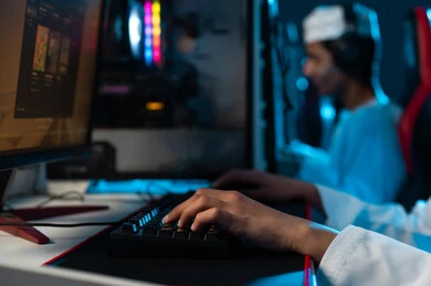 النقر على لوحة مفاتيح الحاسب المكتبي , شاب عربي خليجي عماني يرتدي الدشداشة البيضاء و الكمة المطرزة يجلس على المقعد الجلدي الاحمر , النظر الي شاشة تعرض اللعبة الالكترونية , مشاركة الاصدقاء الاوقات المسلية